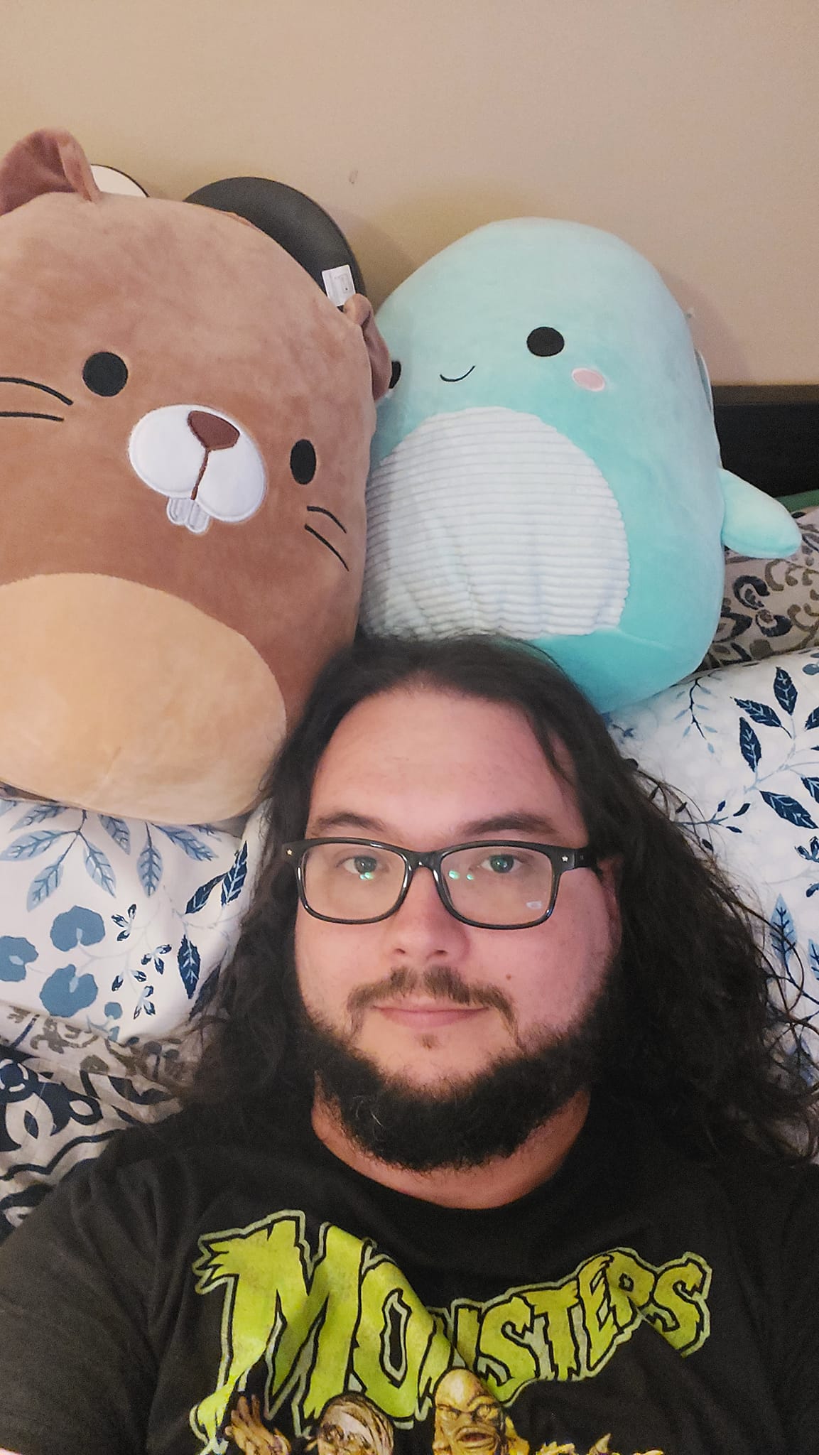 A man with long dark hair and a beard lies in bed with two Squishmallows, a beaver and a Nessie, photo 3